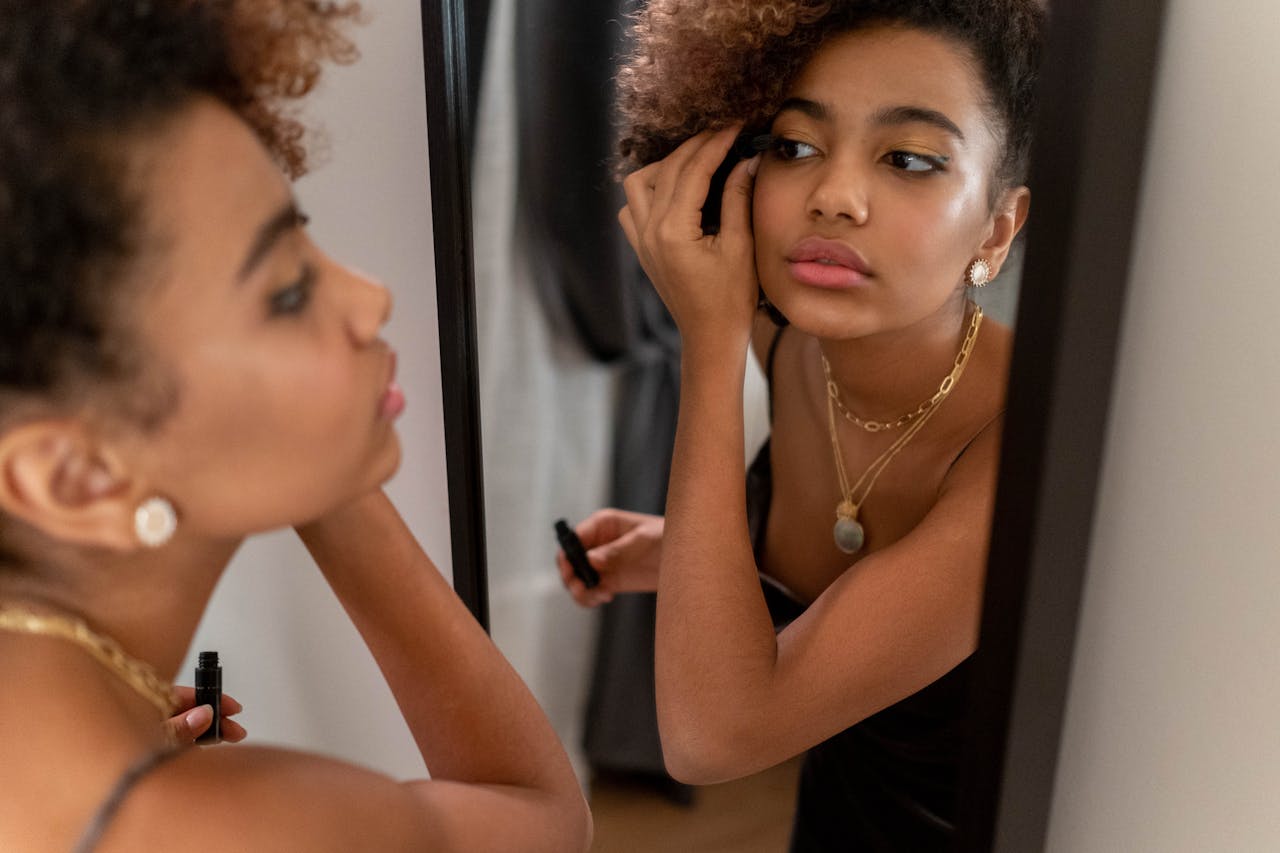A young woman with curly hair applies mascara in front of a mirror, focused on her makeup.